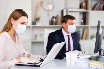 Focused young woman in medical face mask and latex gloves working on laptop in office with male colleague. Concept of prevention of viral infection and social distancing