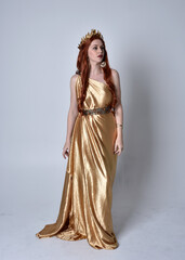Full length portrait of girl with red hair wearing long grecian toga and golden wreath. Standing pose iisolated against a grey studio background. © faestock