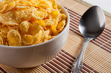 A white bowl of cornflakes, next to a metal spoon. Everything is on a napkin. Horizontal frame.