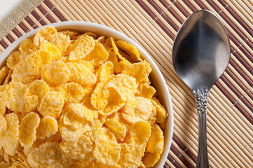 A white bowl of cornflakes, next to a metal spoon. Everything is on a napkin.