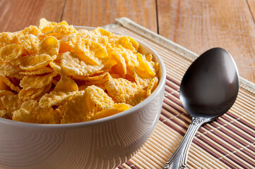 A white bowl of cornflakes, next to a metal spoon, all on a napkin that is on a brown wooden table. Horizontal frame.