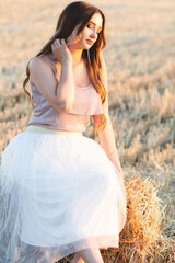 Fototapeta na wymiar Happy woman sitting on hay stack walking in summer evening, beautiful romantic girl with long hair outdoors in field at sunset