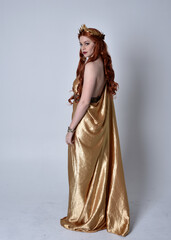 Fototapeta premium Full length portrait of girl with red hair wearing long grecian toga and golden wreath. Standing pose with back to the camera, isolated against a grey studio background.