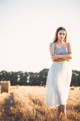 Fototapeta na wymiar young woman in dress walking in evening in field with hay bales, beautiful romantic girl with long hair outdoors in field
