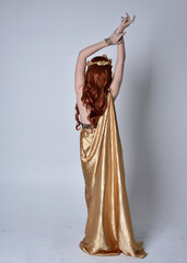 Full length portrait of girl with red hair wearing long grecian toga and golden wreath. Standing pose with back to the camera,  isolated against a grey studio background.