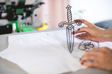 Selective focus of craftswoman holding transparent stencil with sketch near template on surface of screen printing machine 