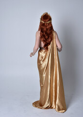 Full length portrait of girl with red hair wearing long grecian toga and golden wreath. Standing pose with back to the camera,  isolated against a grey studio background. - obrazy, fototapety, plakaty