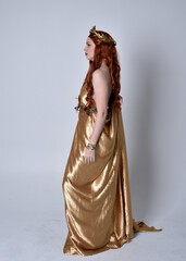 Full length portrait of girl with red hair wearing long grecian toga and golden wreath. Standing pose in side profile,  isolated against a grey studio background.