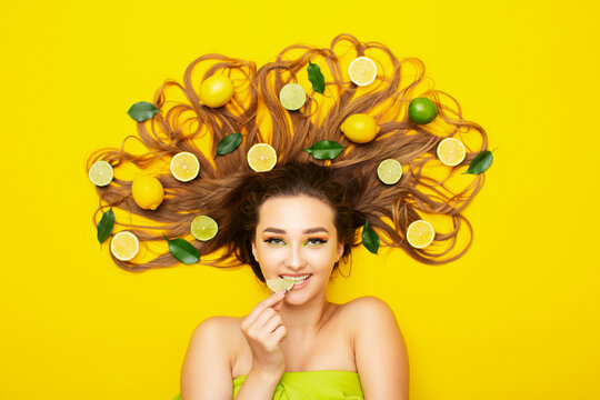 girl lying on yellow background with citrus fruits on long hair,young woman bites lemon wedge, food taste - Powered by Adobe