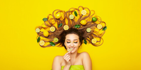 girl lying on yellow background with citrus fruits on long hair,young woman bites lemon wedge, food taste