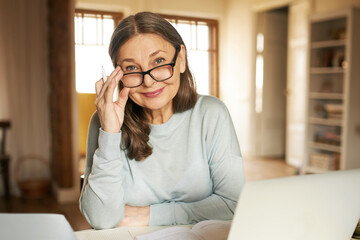 Mature age, people and distant work concept. Attractive confident middle aged woman in glasses working from home, sitting in front of open laptop, doing paperwork, looking at camera with happy smile - Powered by Adobe