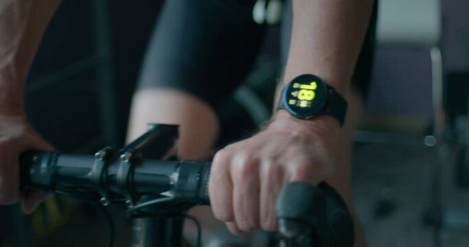 Professional athlete ride indoor bike trainer during high intensity effort workout. Cyclist prepares for summer racing season at home, checks fitness watch to see heart rate stats