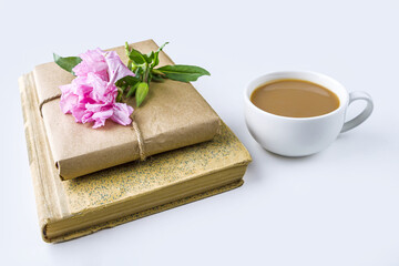 Romantic vintage still life with old book, cup of tea or coffee, pretty gift box wrapped with craft paper and decorated with pink flower on white background.