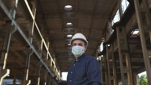 Man, contractor engineer, manager, in blue shirt, protective antibacterial mask came construction site, work, put a white protective helmet on his head. Columns for dismantling in the background