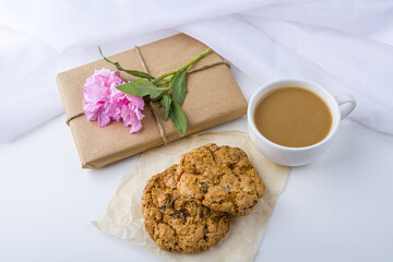 Romantic vintage still life with pretty gift box wrapped with brown craft paper and decorated with pink flower on white background. Dessert (oat cookies) served for tea or coffee break.