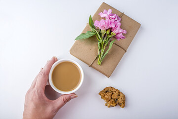 Obraz na płótnie Canvas Romantic vintage still life with pretty gift box wrapped with brown craft paper and decorated with pink flower on white background. Female hand holds cup of coffee