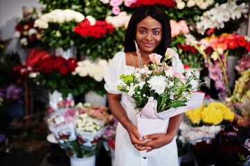 Beautiful african american girl in tender white dress with bouquet flowers in hands standing against floral background in flower shop.Black female florist.