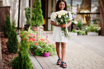 Fototapeta premium Beautiful african american girl holding bouquet of white roses flowers on dating in the city. Black businesswoman with bunch of flowers.