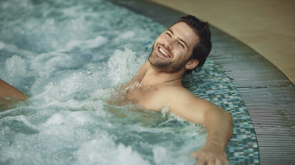 Smiling man relaxing in jacuzzi spa indoor. Sexy guy resting in whirlpool bath - obrazy, fototapety, plakaty