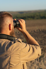 A bearded man looks through binoculars, close-up. The hunter uses binoculars to search