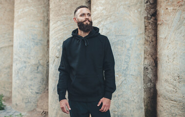 Obraz premium City portrait of handsome hipster guy with beard wearing black blank hoodie or sweatshirt with space for your logo or design. Mockup for print