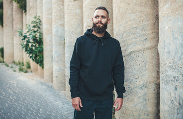 Fototapeta na wymiar City portrait of handsome hipster guy with beard wearing black blank hoodie or sweatshirt with space for your logo or design. Mockup for print