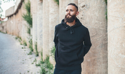 Fototapeta na wymiar City portrait of handsome hipster guy with beard wearing black blank hoodie or sweatshirt with space for your logo or design. Mockup for print