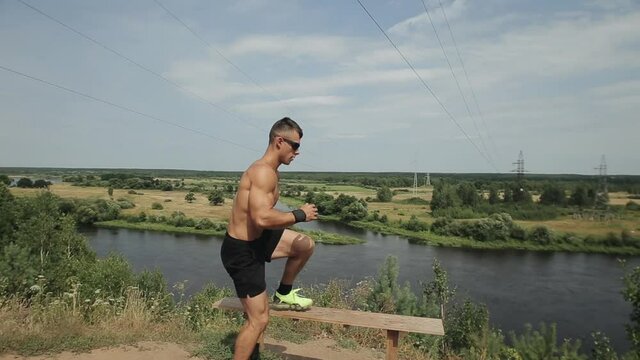 A young bodybuilder plays sports on a sunny day by the river and does exercises on the muscles of the legs. Camera moves up and down
