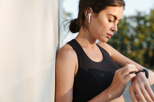 Image of focused athletic sportswoman in headphones using smartwatch