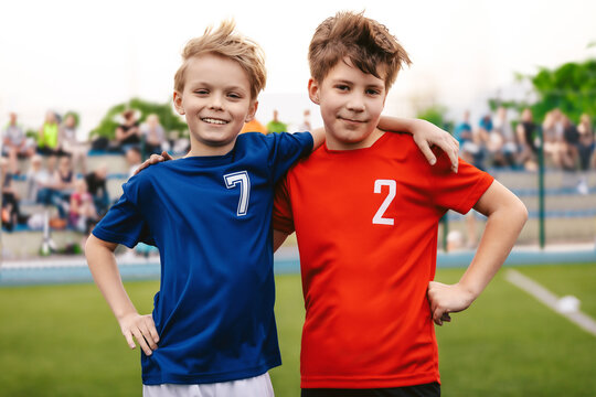 Two Happy Smiling Boys in Sports Team Standing on Grass Pitch. Kids Making Sports Activity Outdoor. Children in Sports Jersey Uniforms. Spectators and Parents in Blurred Background