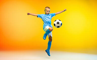 Fototapeta na wymiar In flight. Young boy as a soccer or football player in sportwear practicing on gradient yellow studio background in neon light. Fit playing boy in action, movement, motion at game. Copyspace.