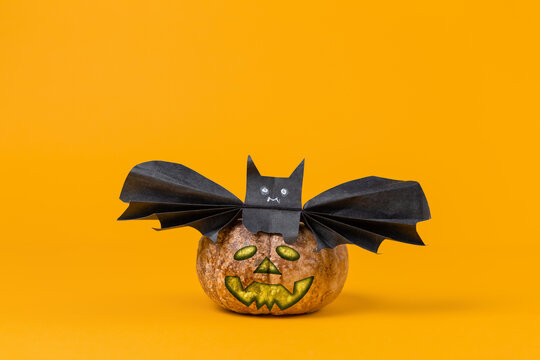 A pumpkin with an image of a face, on which a bat is sitting, made of paper. Orange background. Copy space. The concept of Halloween, and holiday decorations