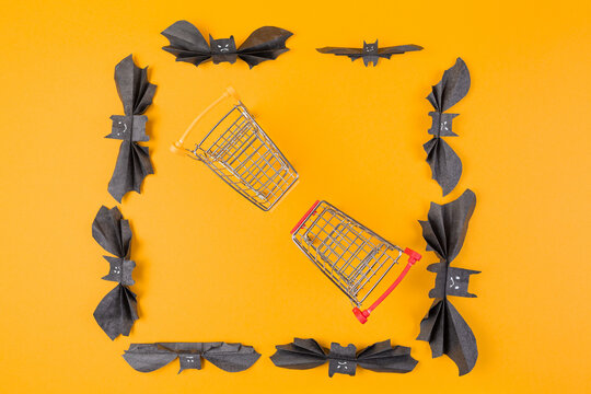 Two shopping toy carts are located in a frame of paper decorative bats. Orange background. Copy space. Flat lay. The concept of Halloween and holiday shopping