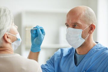Modern doctor wearing mask sitting in front of senior patient taking nasal swab test for coronavirus