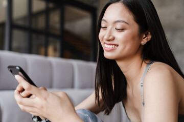 Image of happy asian woman using mobile phone while sitting on floor