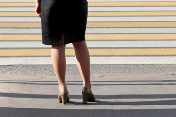 Woman in black leather skirt standing in front of a crosswalk, rear view. Female legs in shoes on on a street, concept of road safety