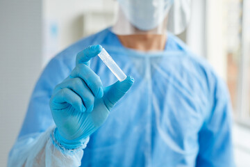 Horizontal medium close-up shot of unrecognizable medical worker wearing protective uniform holding ampoule with transparent liquid