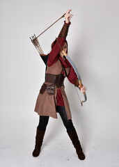 Full length portrait of girl with red hair wearing  brown medieval archer costume.. Standing pose holding a bow and arrow,  isolated against a grey studio background.