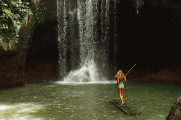 Rear view of young woman standing in front of waterfall on bamboo raft