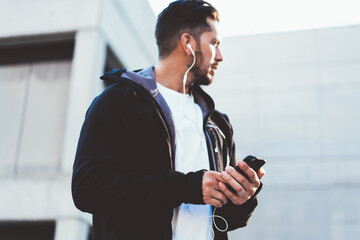 Rear view of sportsman in trendy jacket and earphones holding mobile phone standing on urban settings, male in trendy active outfit using technology for listening online radio during workout on street