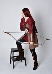 Full length portrait of girl with red hair wearing  brown medieval archer costume.. Standing pose holding a bow and arrow,  isolated against a grey studio background.