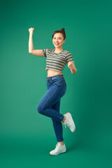 Full length portrait of happy young Asian woman standing on one leg and dancing on green background.