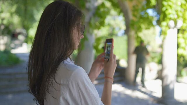 Tourist woman taking photos of beautiful green park and ancient buildings using her smartphone