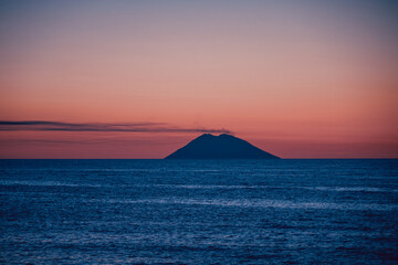 Naklejka na ściany i meble Amazing view of the Stromboli Volcano at sunset, wit the sun going down and amazing sea and sky colors. View form the coast of Calabria, Italy, in the touristic destination of Tropea.