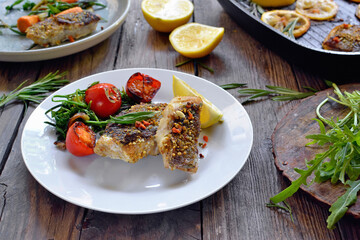 Grilled fish with lemon, herbs and rosemary. Fried fish fillets in a white plate. Beautiful presentation of food. Rucolla salad and carrots. Wooden background