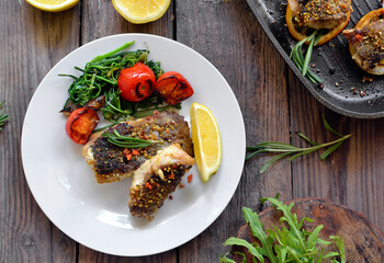 Grilled fish with lemon, herbs and rosemary. Fried fish fillets in a white plate. Beautiful presentation of food. Rucolla salad and carrots. Top view. Wooden background