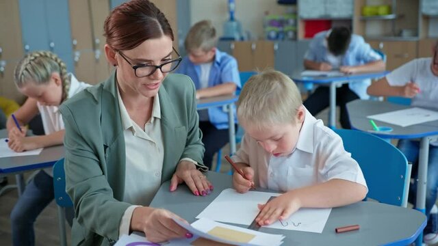 disabled boy with down syndrome studys shapes with teacher sitting at desk on the background of classmates during the lesson at school