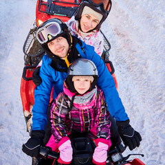 Portrait of happy family of three, wearing colorful ski suits, helmets, spending winter holiday together in snowy mountains on red quad bike, top view. Concept of active leisure and quad biking.