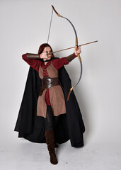 Full length portrait of girl with red hair wearing medieval archer costume with black cloak. Standing pose with back to the camera holding a bow and arrow,  isolated against a grey studio background.