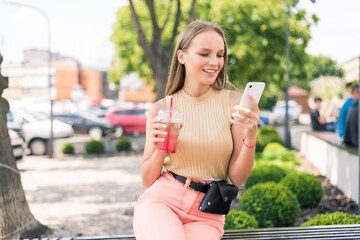 Young woman with phone chating or surf in internet drinks mojito fruit cocktail in the street